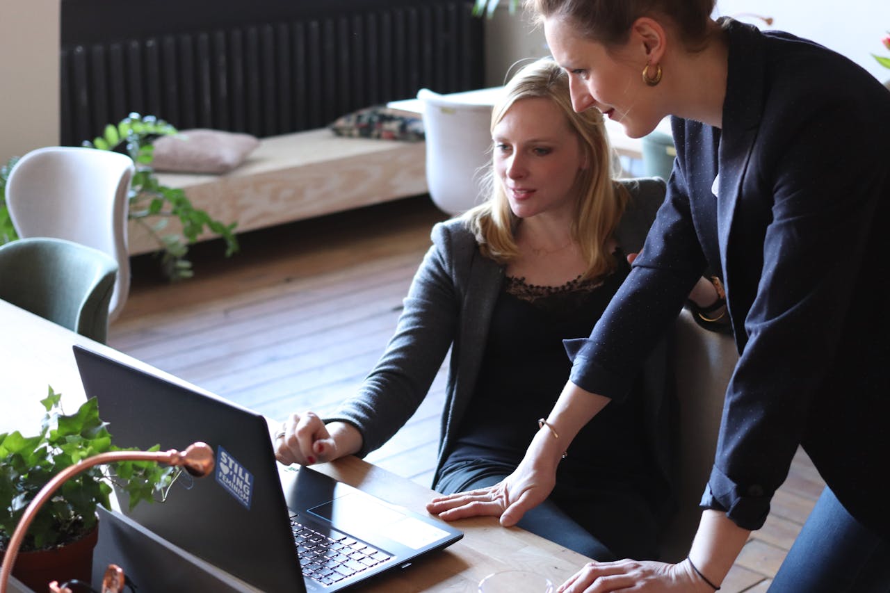 about-04 Two professional women discuss a project using a laptop in a modern office environment.