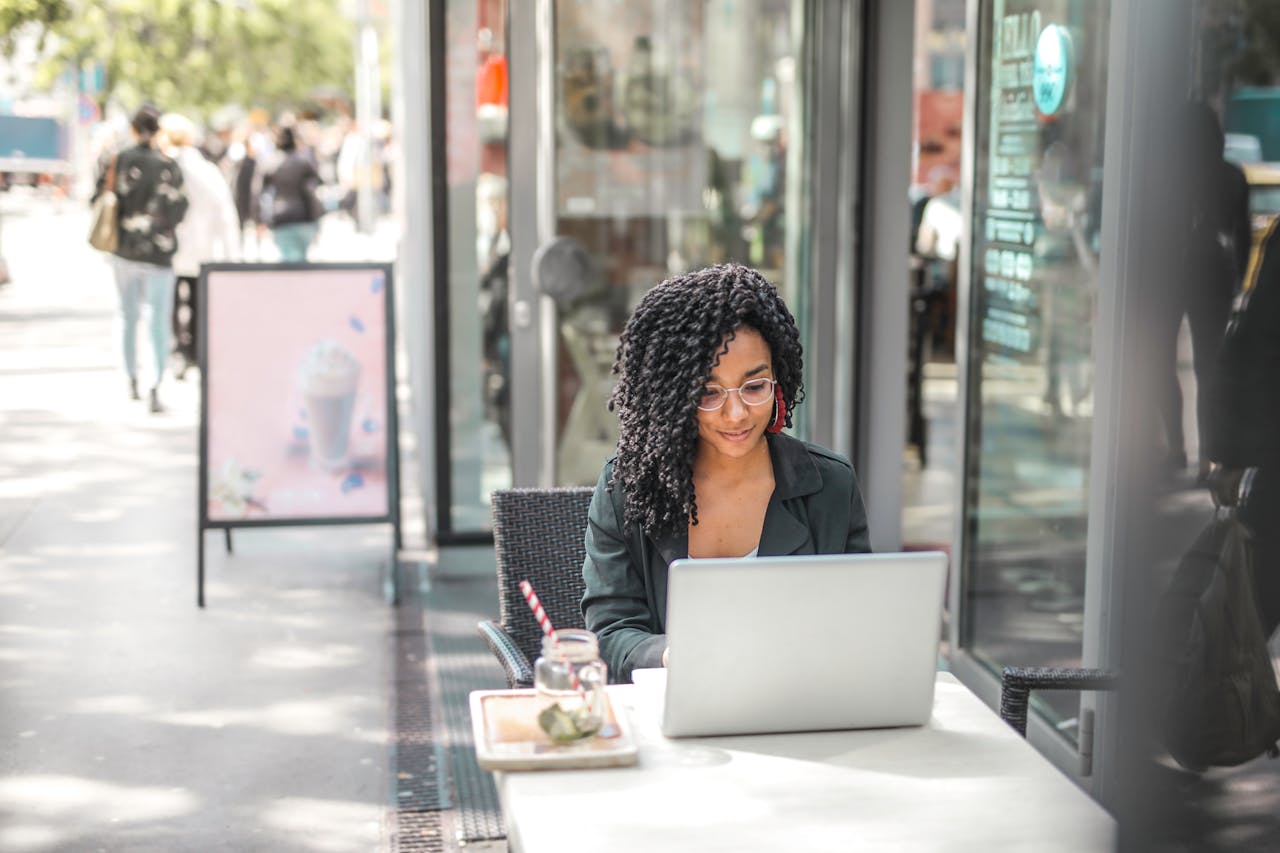 Home High angle of pensive African American female freelancer in glasses and casual clothes focusing on screen and interacting with netbook while sitting at table with glass of yummy drink on cafe terrace in sunny day