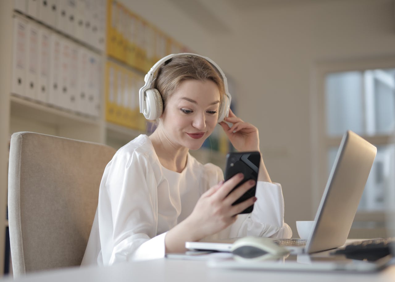 about-002 Woman with headphones smiling at smartphone while working on laptop indoors