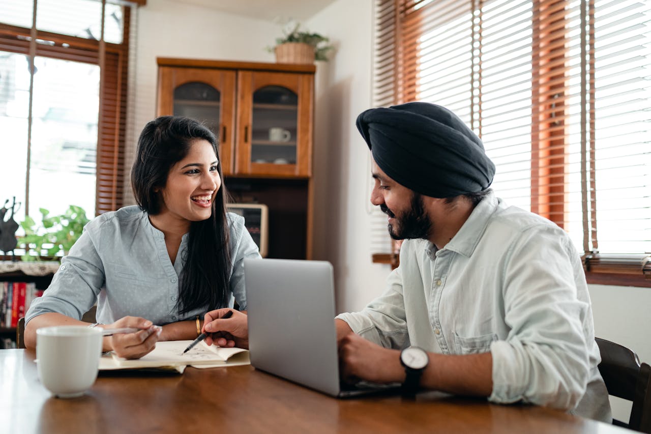 Home Laughing Indian woman with husband sitting at table and discussing project while working together with laptop and notebook at home